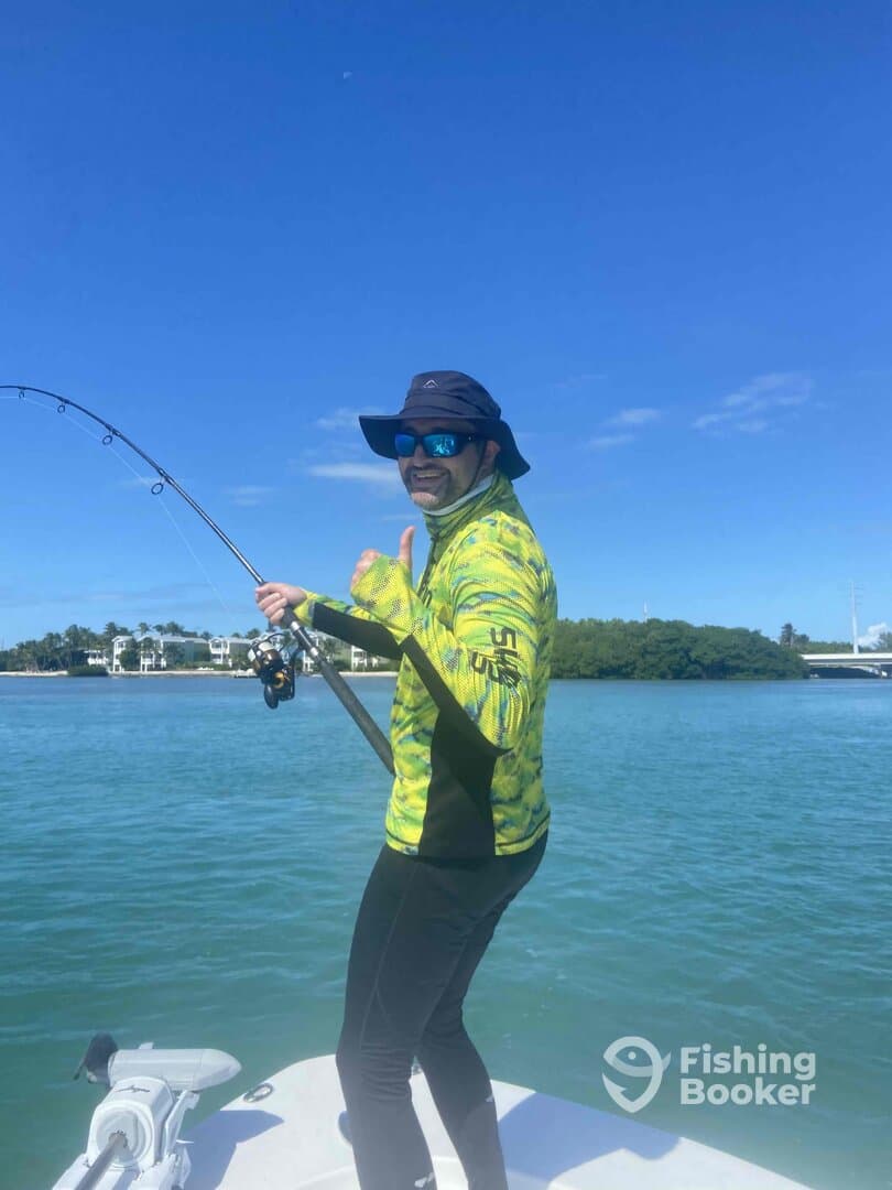 Angler reeling in a catch while standing on the bow of a boat in a sunny coastal environment.