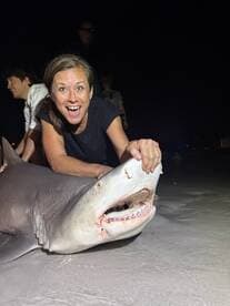 An excited angler poses with a large shark on the beach at night, showcasing a thrilling catch during a night fishing adventure.