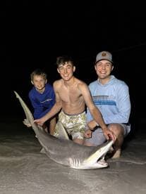 A group of young anglers proudly displaying a large shark caught during a nighttime fishing adventure on the beach.