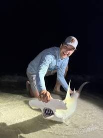 An enthusiastic angler proudly displaying a Hammerhead Shark while wading in the shallow waters at night.
