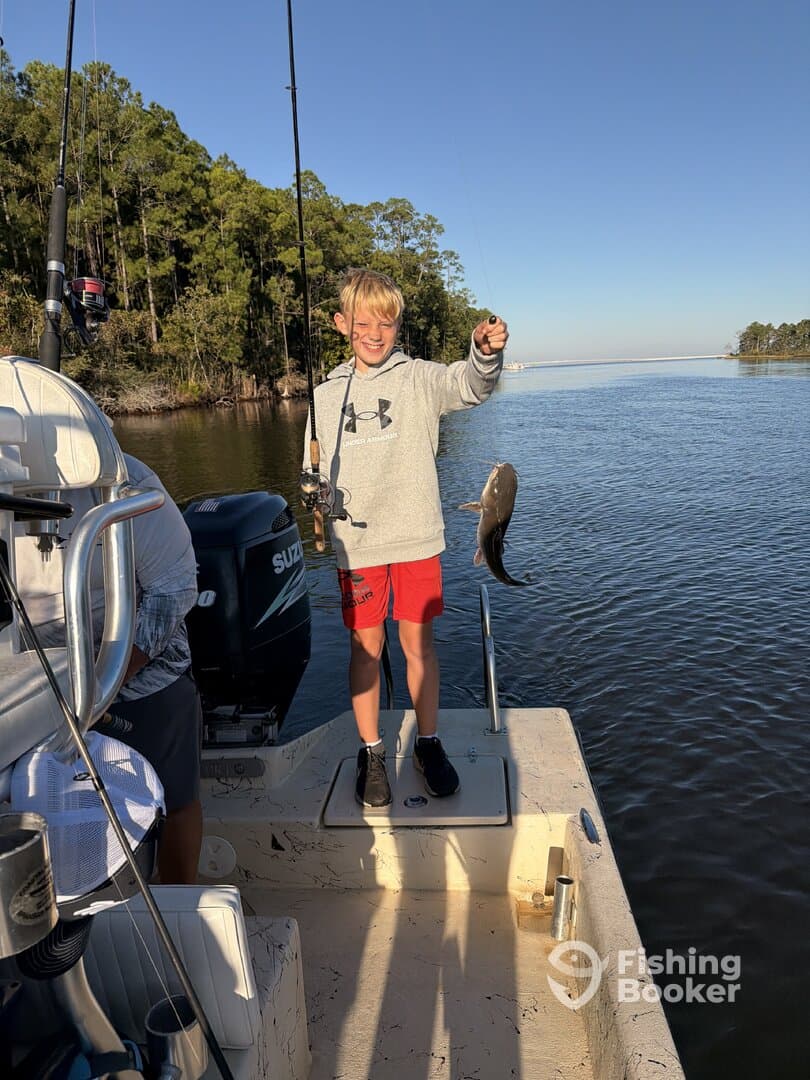 A young angler joyfully displaying a freshly caught fish while fishing from a boat in a serene waterway surrounded by lush greenery.
