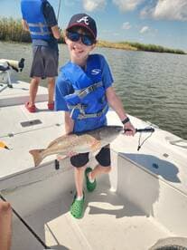 A young angler proudly displaying a large Redfish while fishing on a sunny day in a scenic waterway.