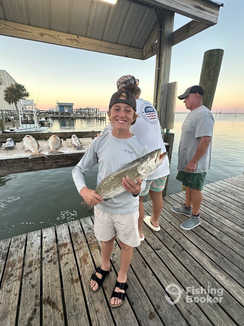 A young angler proudly displaying a large Speckled Trout on the dock after a successful fishing trip.