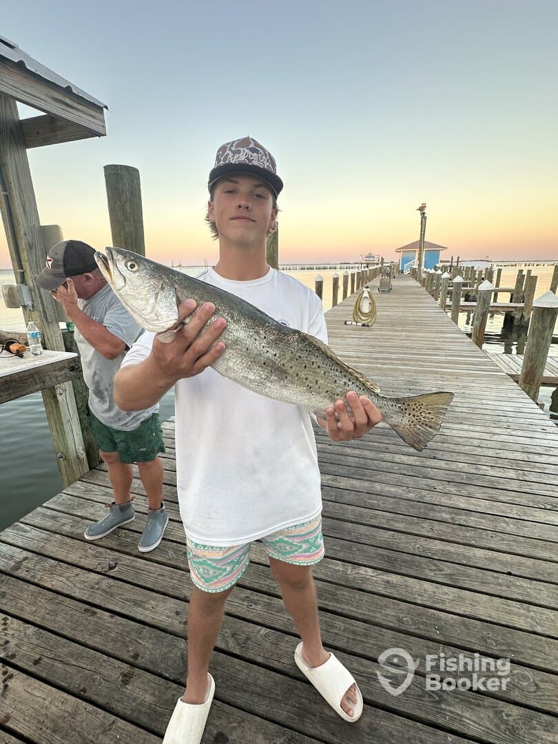 A young angler proudly displaying a large Speckled Trout on a dock during a sunset fishing trip.