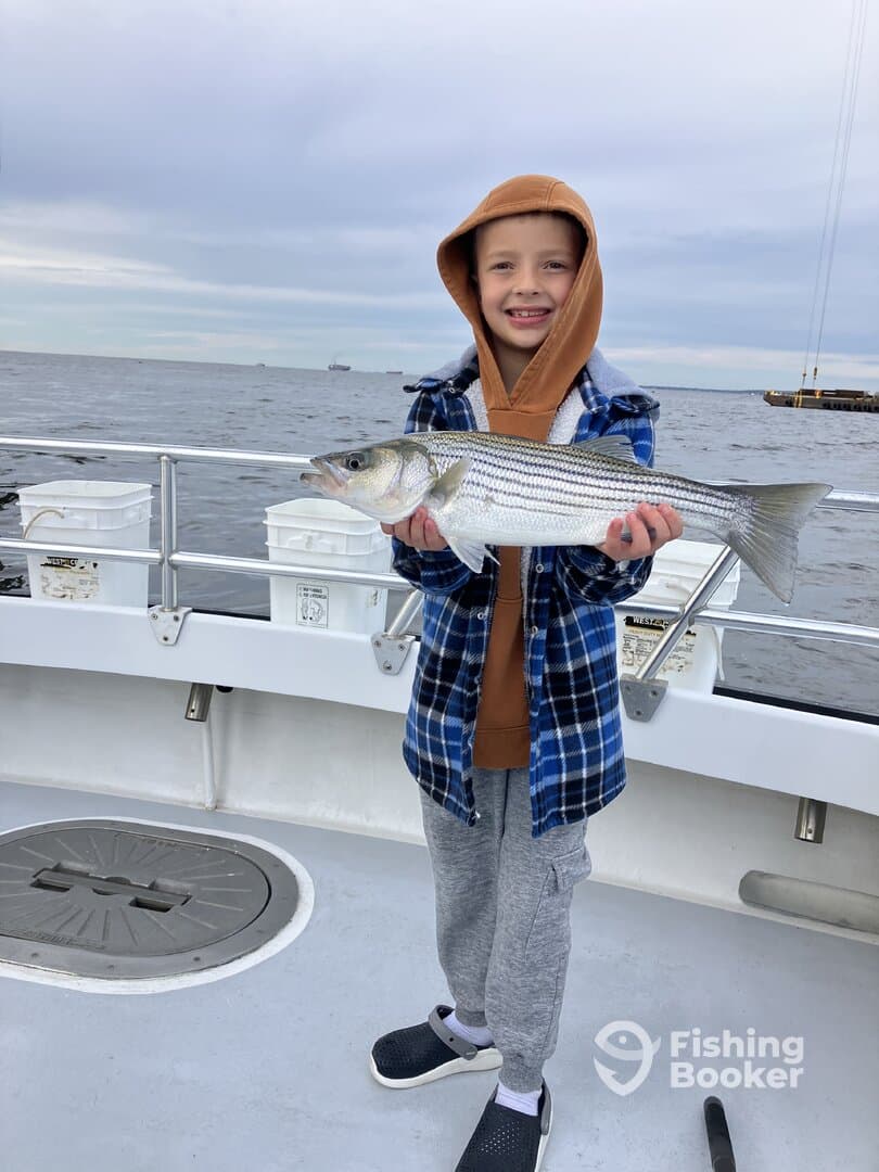 A young angler proudly displaying a striped bass while fishing on a boat, showcasing a successful outing.