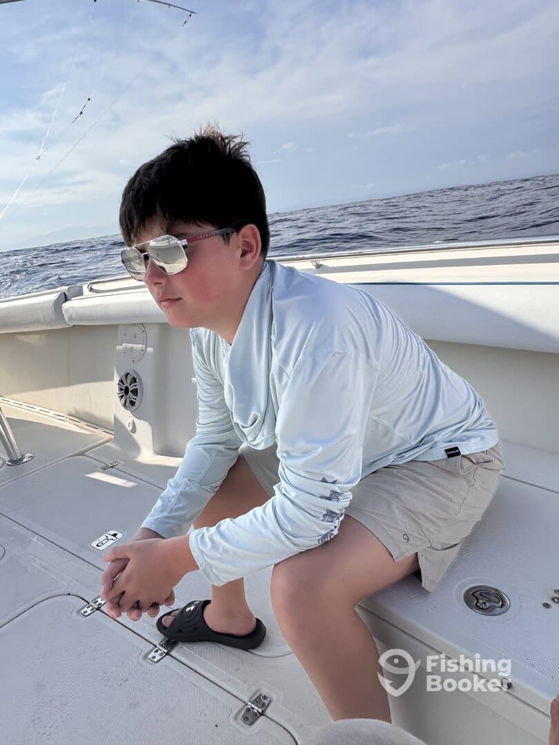A young boy seated on the deck of a fishing boat, wearing sunglasses and a long-sleeve shirt, enjoying the ocean view.