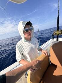 A young angler on a fishing boat, wearing sunglasses and a hoodie, appears focused while enjoying a day on the open water.