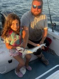 A young girl proudly holds a small shark while sitting on a boat, showcasing a memorable fishing experience with her companion.
