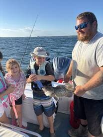 A young boy proudly holds a caught fish with the help of an adult, while two girls look on excitedly during a family fishing trip.