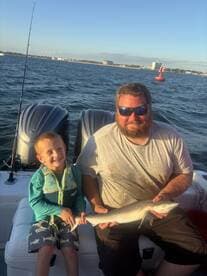 A young boy and an adult proudly display a caught shark while enjoying a fishing trip on a boat, showcasing a beautiful coastal backdrop.