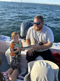A young boy proudly holding a small shark while fishing with an adult on a boat, showcasing a fun family fishing experience.