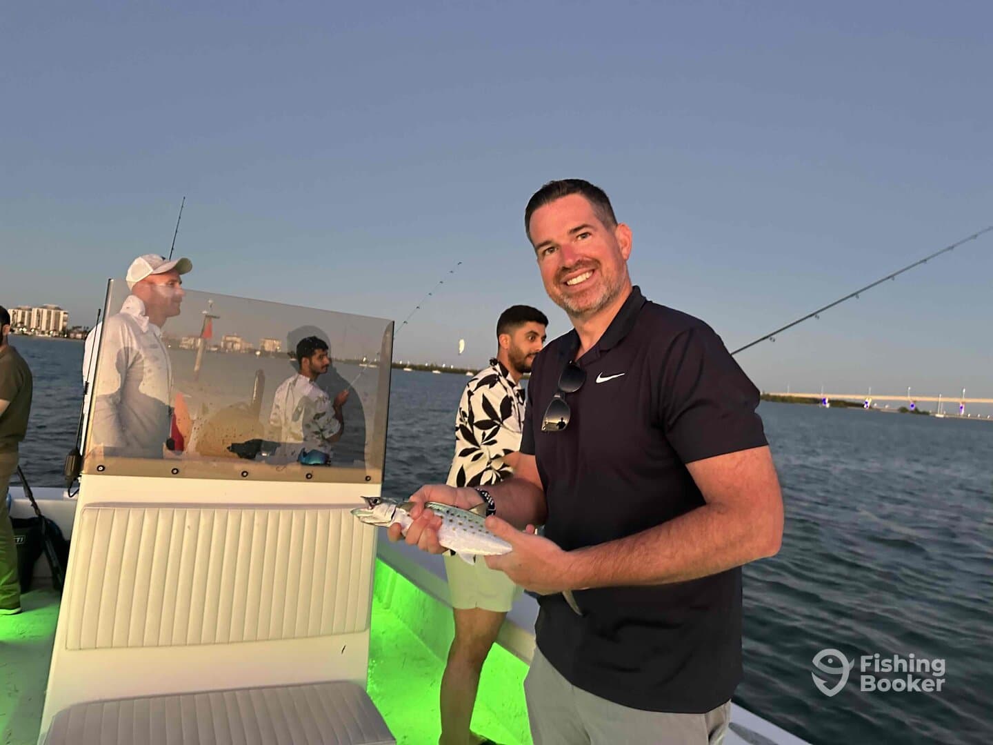 An angler proudly displaying a caught fish while enjoying a sunset fishing trip on a boat, with friends in the background.