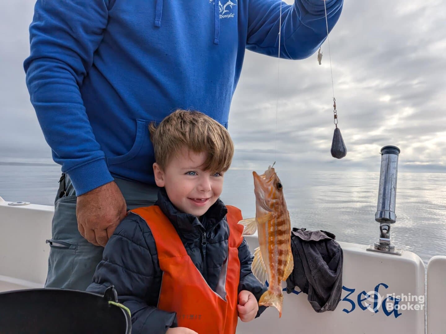 A young boy in an orange life jacket beams with joy as he holds up a freshly caught fish while fishing on a boat.
