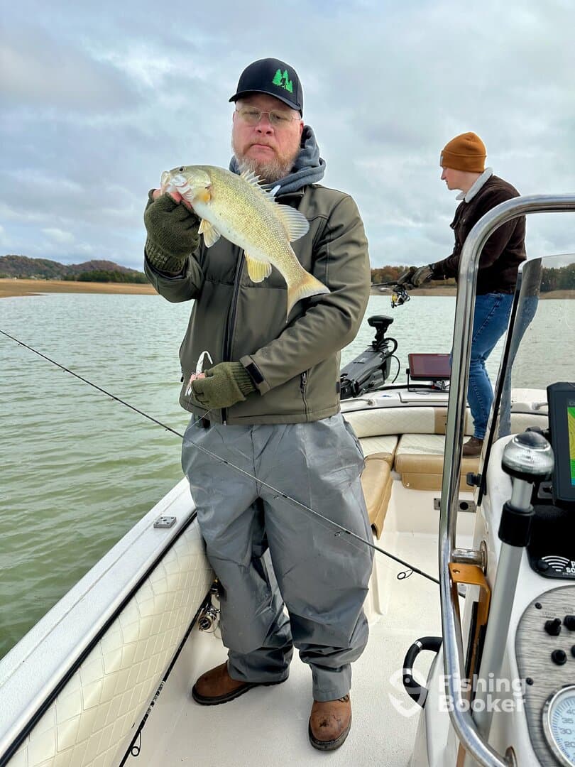 An angler proudly displaying a large Largemouth Bass while fishing on a lake, with another person in the background preparing to cast.