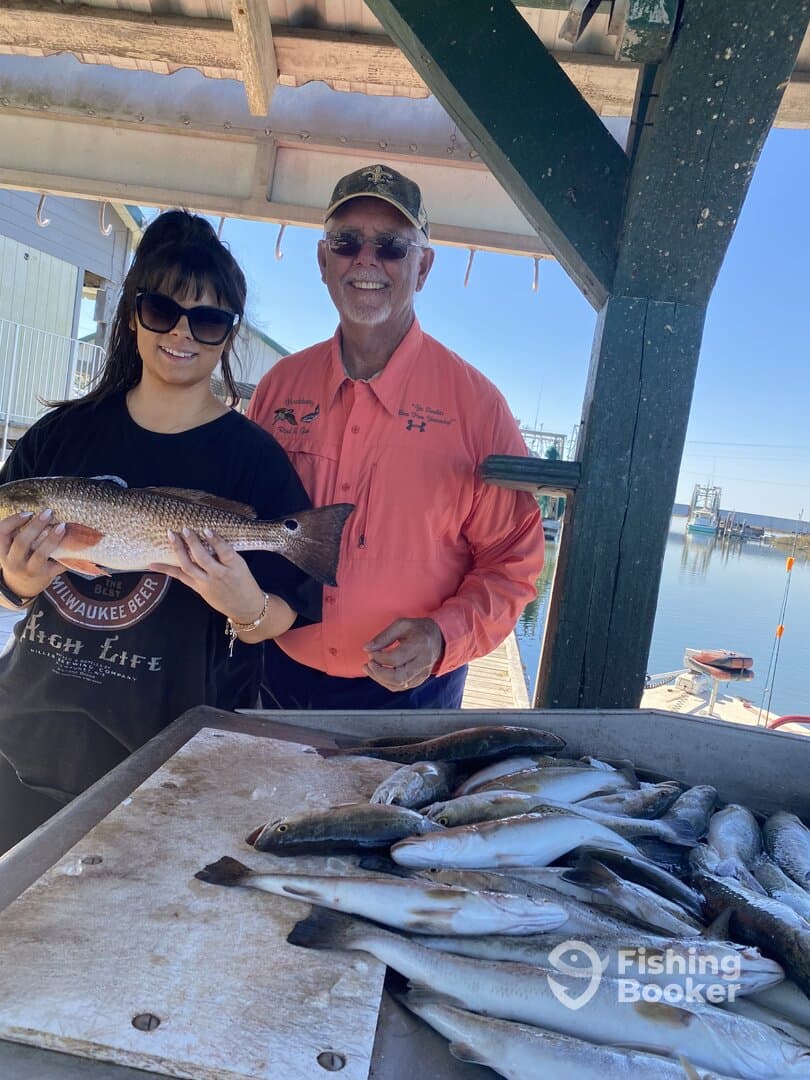 A young angler and an older gentleman proudly displaying a large Speckled Trout at the dock, surrounded by a variety of freshly caught fish.