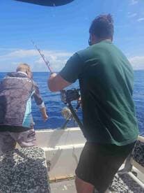 Two anglers engaged in fishing on a boat, with one reeling in a line while the other prepares to assist, showcasing a vibrant blue ocean backdrop.