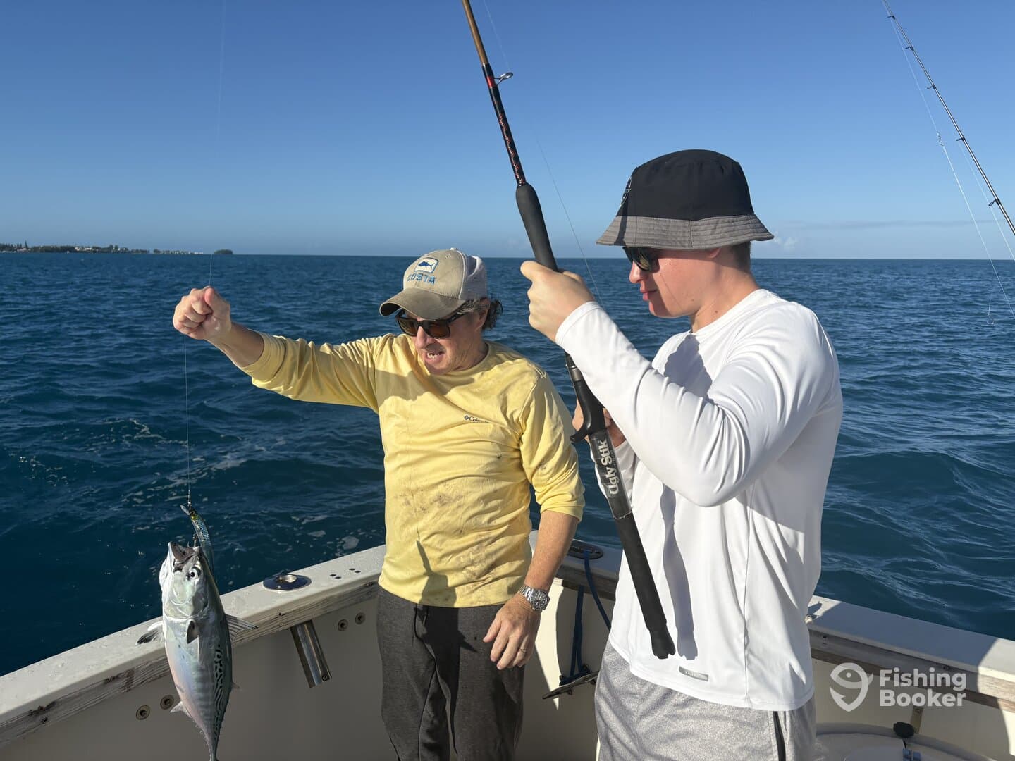 Two anglers actively fishing on a boat, with one holding a caught fish, likely a Mackerel, while the other prepares to reel in.