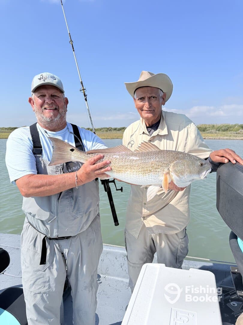 Two anglers proudly displaying a large Redfish on a boat during a fishing trip in a coastal environment.