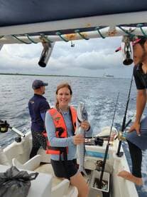 A woman in a life jacket proudly displaying a Barracuda while fishing on a boat, with fellow anglers visible in the background.