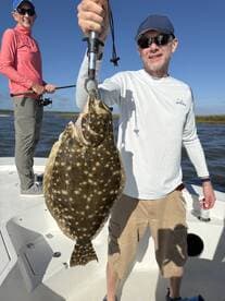 An angler proudly displays a large Flounder while fishing on a sunny day, with another person in the background preparing to cast.