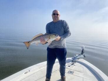 An angler proudly displaying a large Redfish while standing on the bow of a boat in calm waters.
