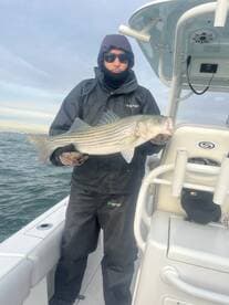 Angler displaying a large Striped Bass while aboard a boat, showcasing a successful fishing trip in cooler waters.