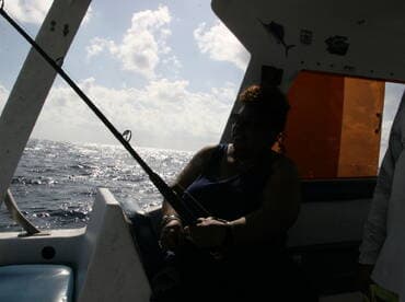 An angler engaged in fishing on a boat, casting a line into the open ocean under a bright sky.