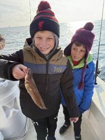 Two young anglers smiling while holding a small fish on a boat, enjoying a fishing trip.
