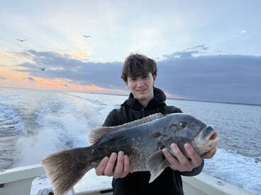 A young angler proudly displaying a large Black Sea Bass while on a boat, with a beautiful sunset backdrop over the water.