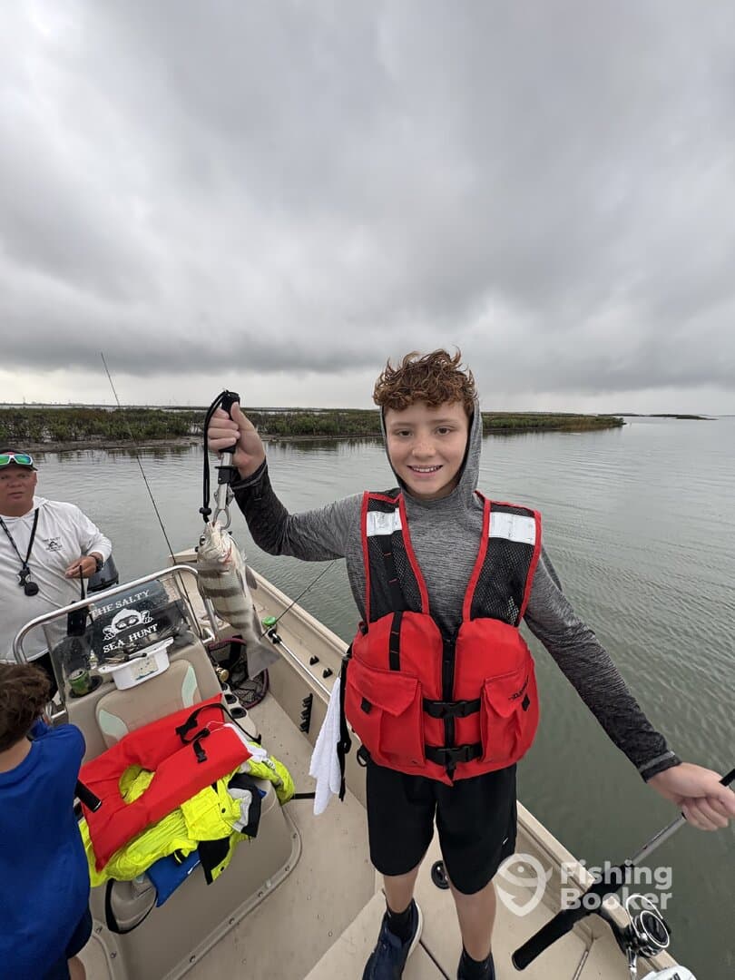 A young angler proudly displaying a striped bass while wearing a life jacket on a fishing boat in a coastal environment.