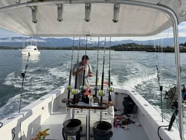 A fisherman prepares gear on the deck of a sportfishing boat, with scenic mountains in the background and a second boat in the distance.