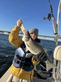 A young angler proudly displaying a striped bass while fishing from a boat, with a scenic bridge in the background.