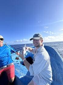 Two anglers on a small boat enjoying a successful fishing trip, with one proudly displaying a small shark against a backdrop of clear blue skies.