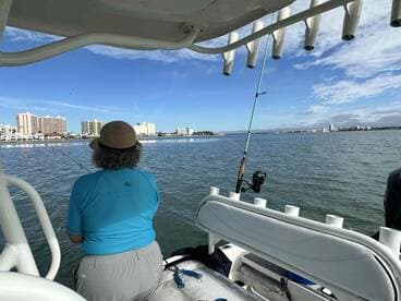 An angler fishing from a boat in a scenic coastal area, with city buildings in the background and a clear blue sky.