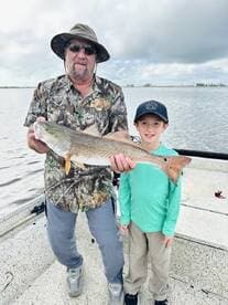 A proud angler and a young boy display a large Redfish on a fishing trip, showcasing a successful catch in a scenic waterway.