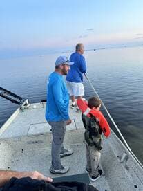 A young boy in a life jacket watches intently as two adults fish from a boat in calm waters during a serene fishing trip.