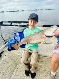 A young angler proudly displaying a large Redfish while sitting on a boat, showcasing a successful fishing trip.