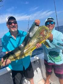 Two anglers proudly displaying a large Mahi Mahi while fishing offshore, showcasing a vibrant blue sky and ocean backdrop.