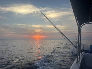 A serene sunset view from the side of a fishing boat, showcasing the calm waters and fishing rods ready for action.