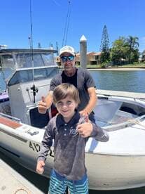 A young boy and an adult are giving thumbs up in front of a fishing boat, showcasing a cheerful moment at the dock.