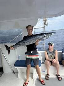 A young angler proudly displaying a large Wahoo on a fishing charter, with family members visible in the background.