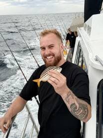 A smiling angler proudly displays a small striped fish while fishing on a boat in open waters.