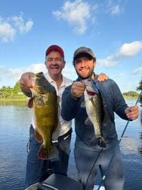 Two anglers proudly displaying their catches, a large Largemouth Bass and a Peacocks Bass, while fishing on a serene lake.