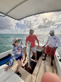 A young angler proudly displays a large Barracuda while fishing with family on a boat in clear waters.