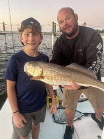 A young angler and an adult proudly displaying a large Redfish on a boat, showcasing a successful fishing trip.