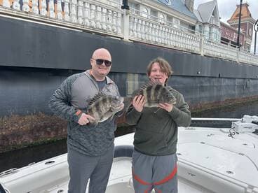 A proud moment for a young angler and an adult, both displaying their catches of striped bass while standing on a boat near a scenic waterfront.
