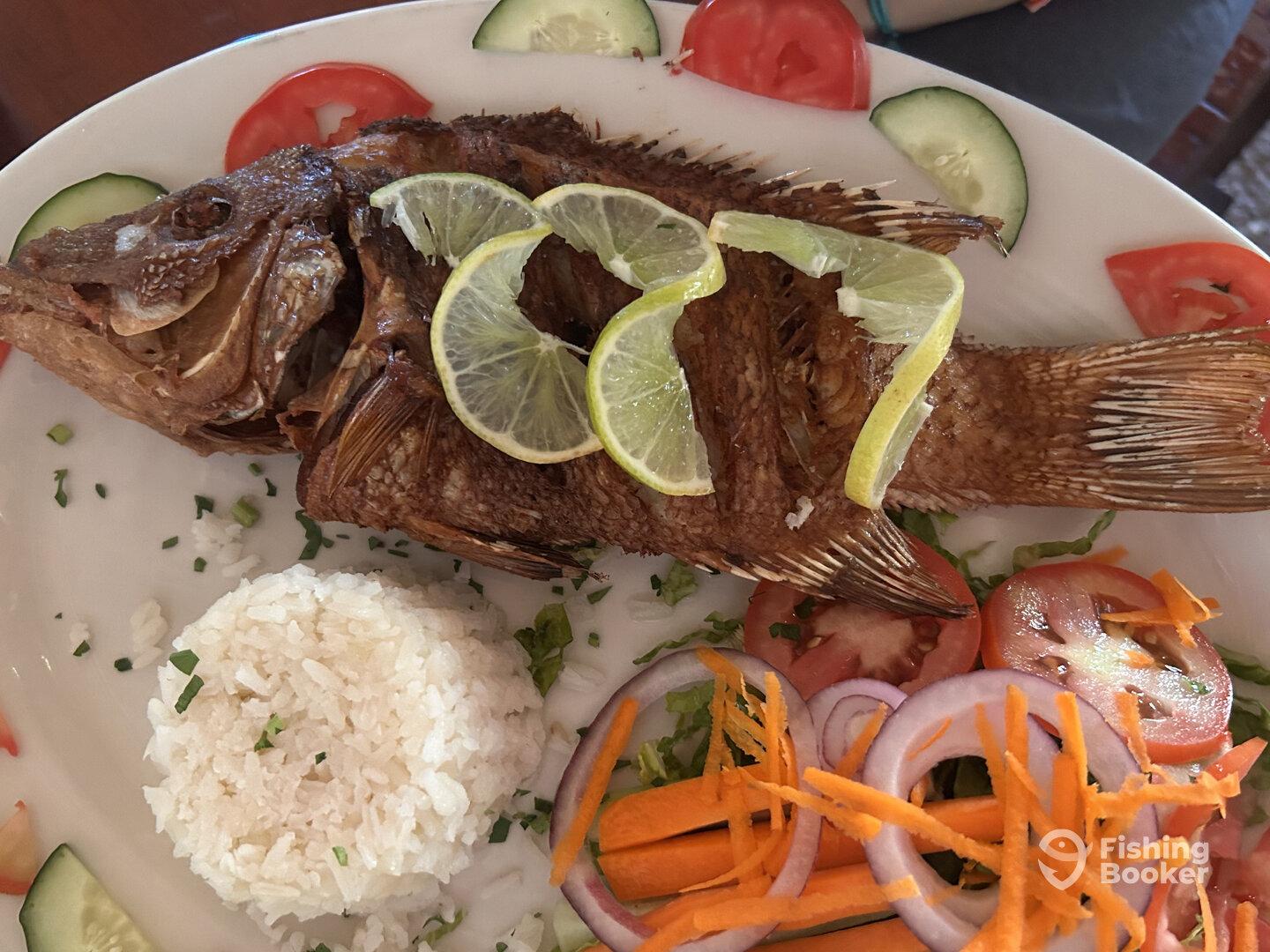 A beautifully plated fish dish featuring a whole fried fish garnished with lime, served with rice and fresh vegetables.