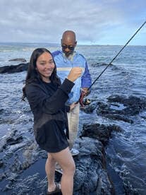 A young angler is smiling while holding a fishing line, with an adult assisting her on rocky shores, showcasing a fun fishing experience.