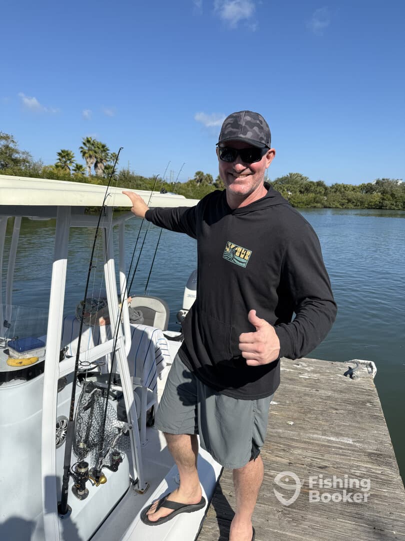 A smiling angler giving a thumbs up next to a fishing boat at the dock, showcasing a sunny day on the water.