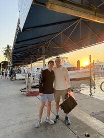 A father and son pose together at the marina during sunset, with boats in the background, capturing a memorable moment after a fishing trip.